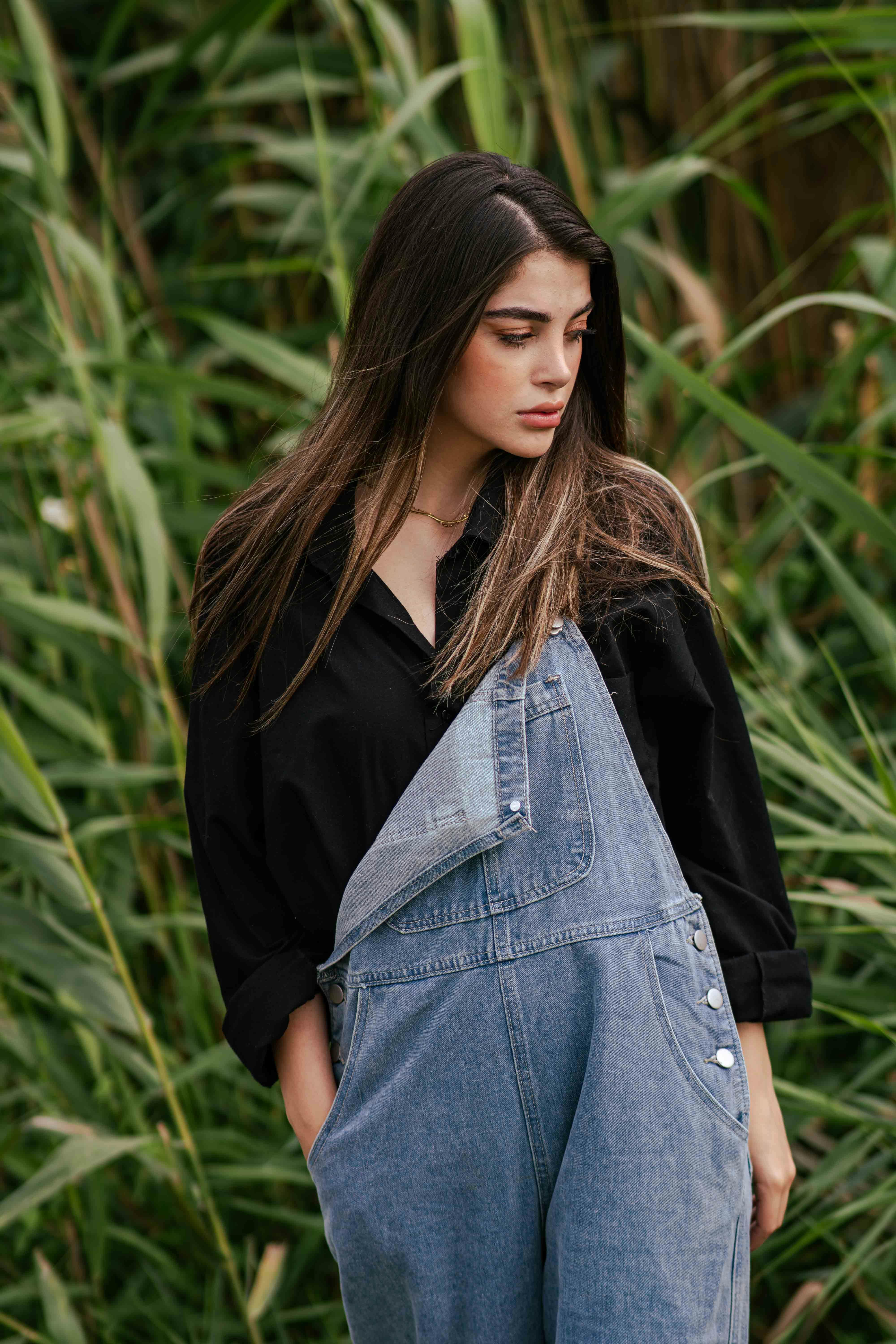 Stylistic outdoor portrait of a woman in denim overalls, amidst lush greenery.