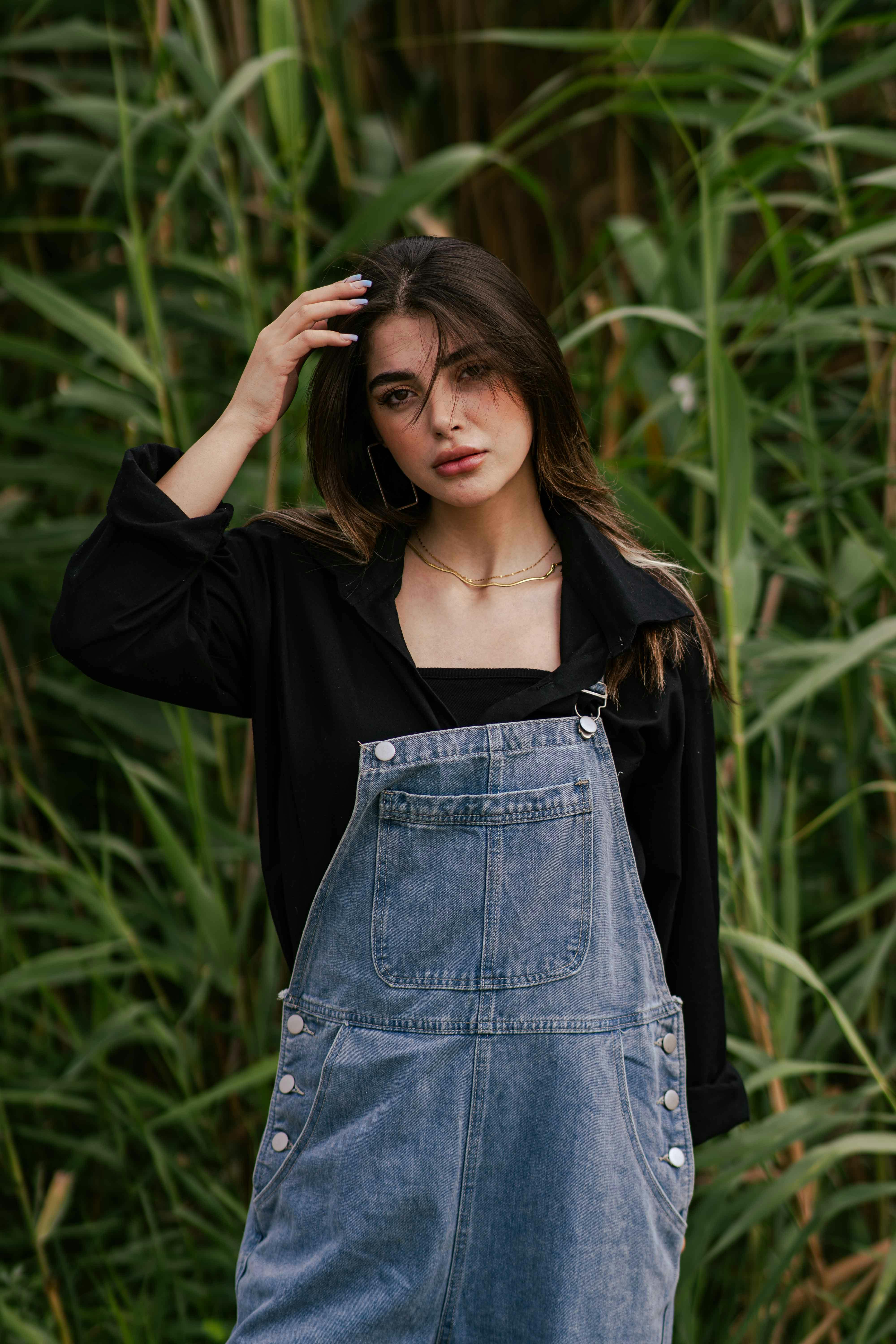 Portrait of a young woman in denim overalls surrounded by lush green foliage.