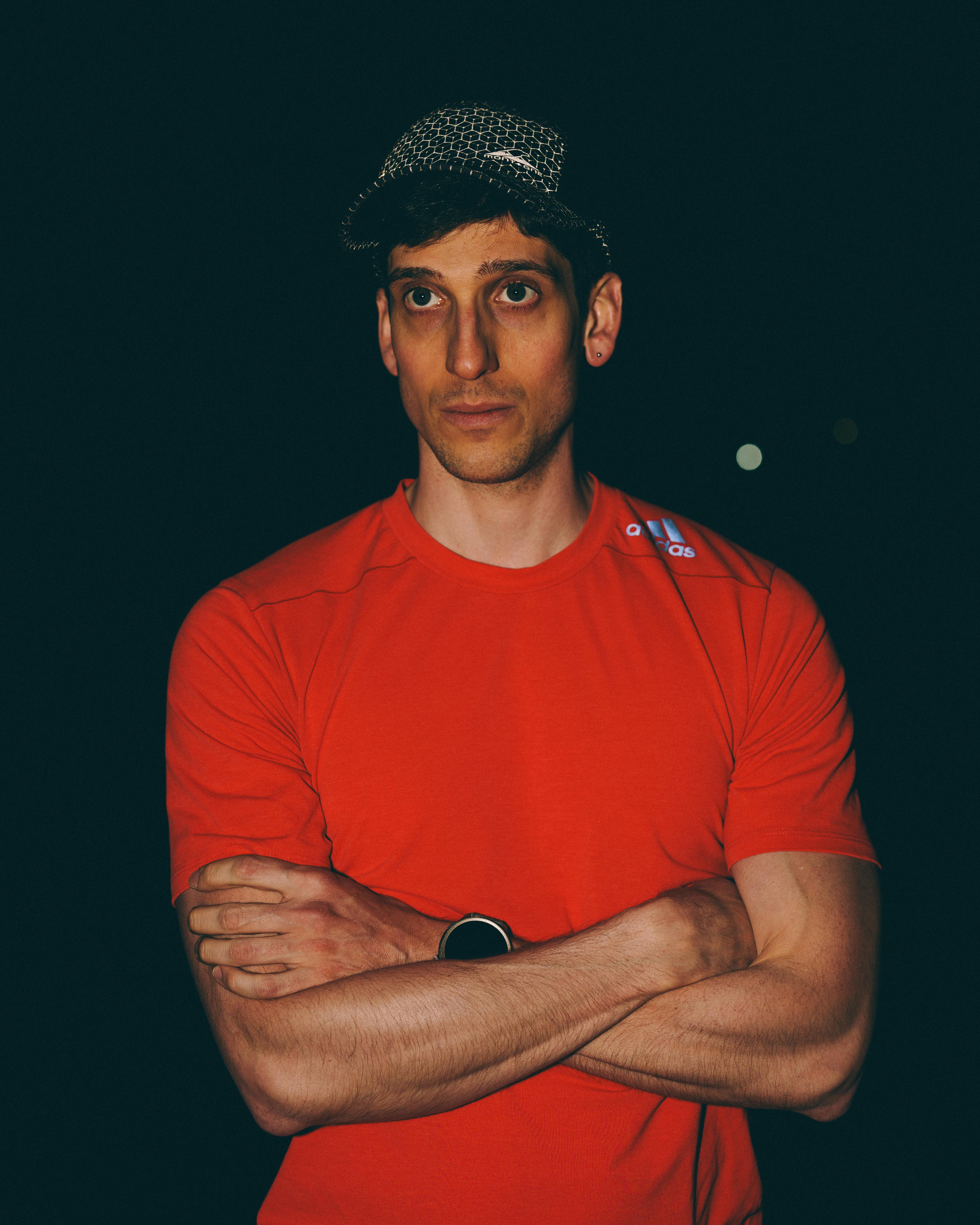 A man in a red tee stands with arms crossed in a night setting.