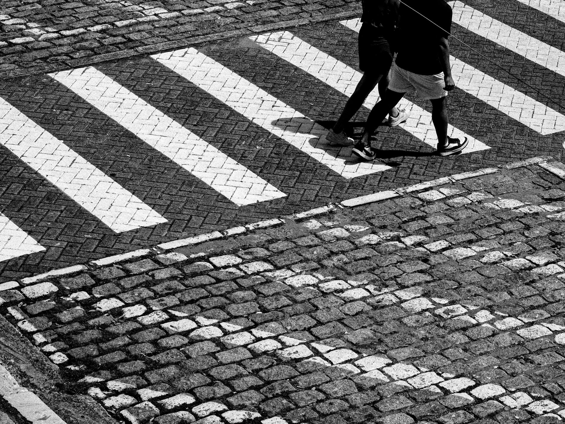 A dynamic black and white photo capturing two pedestrians crossing a cobblestone street, emphasizing patterns and contrasts.