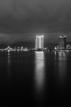 Black and white city skyline at night with illuminated buildings reflecting on the water.