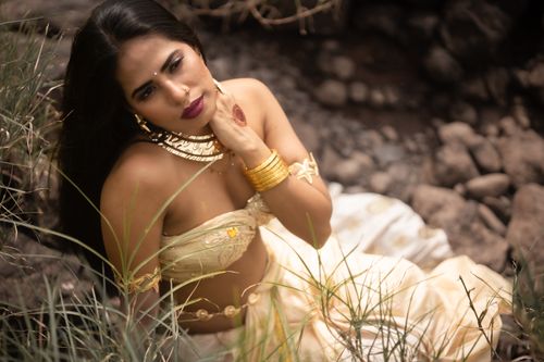 Free A woman in traditional attire sitting outdoors, adorned with gold jewelry and surrounded by nature. Stock Photo