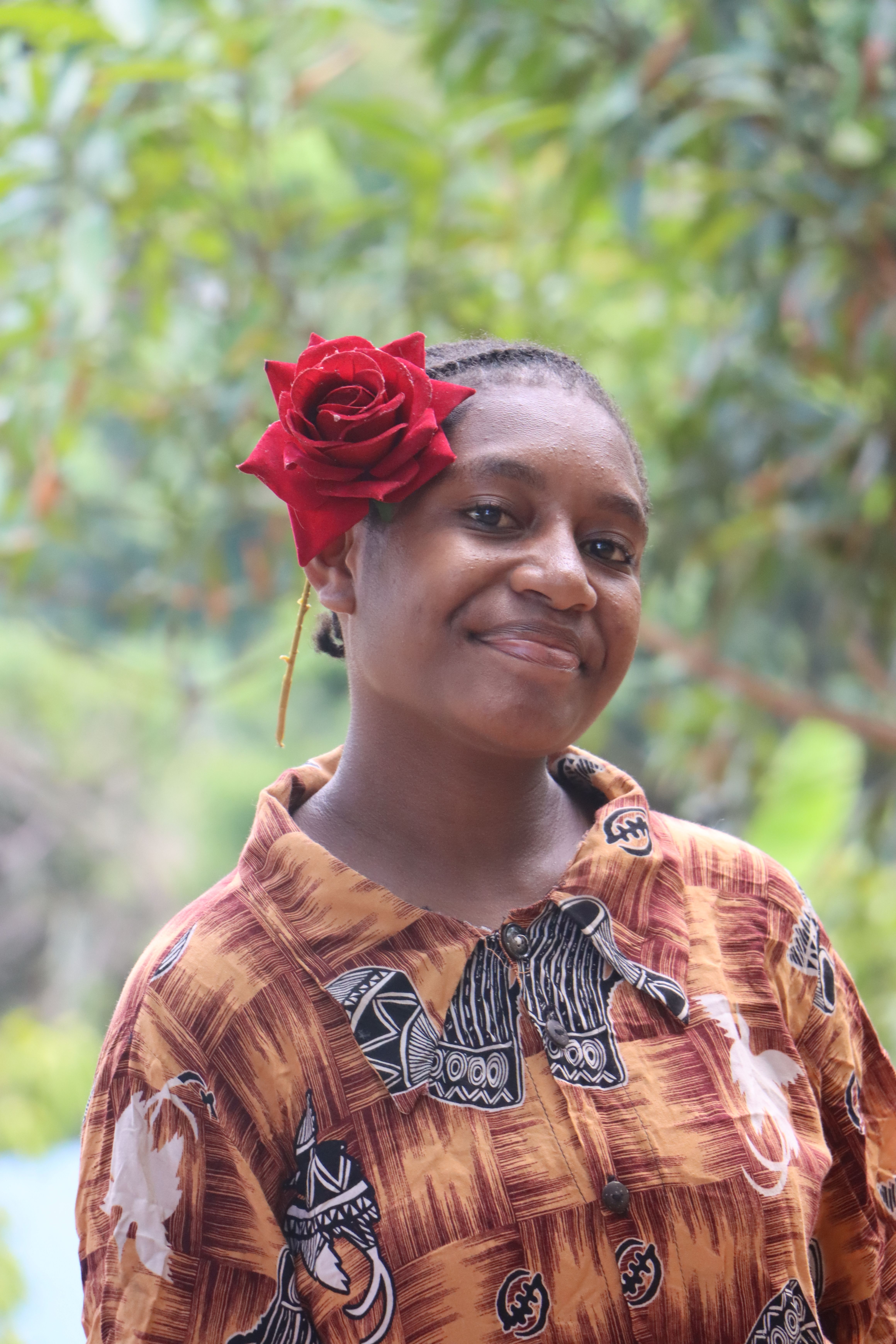 Smiling woman with rose in hair, wearing patterned shirt outdoors.