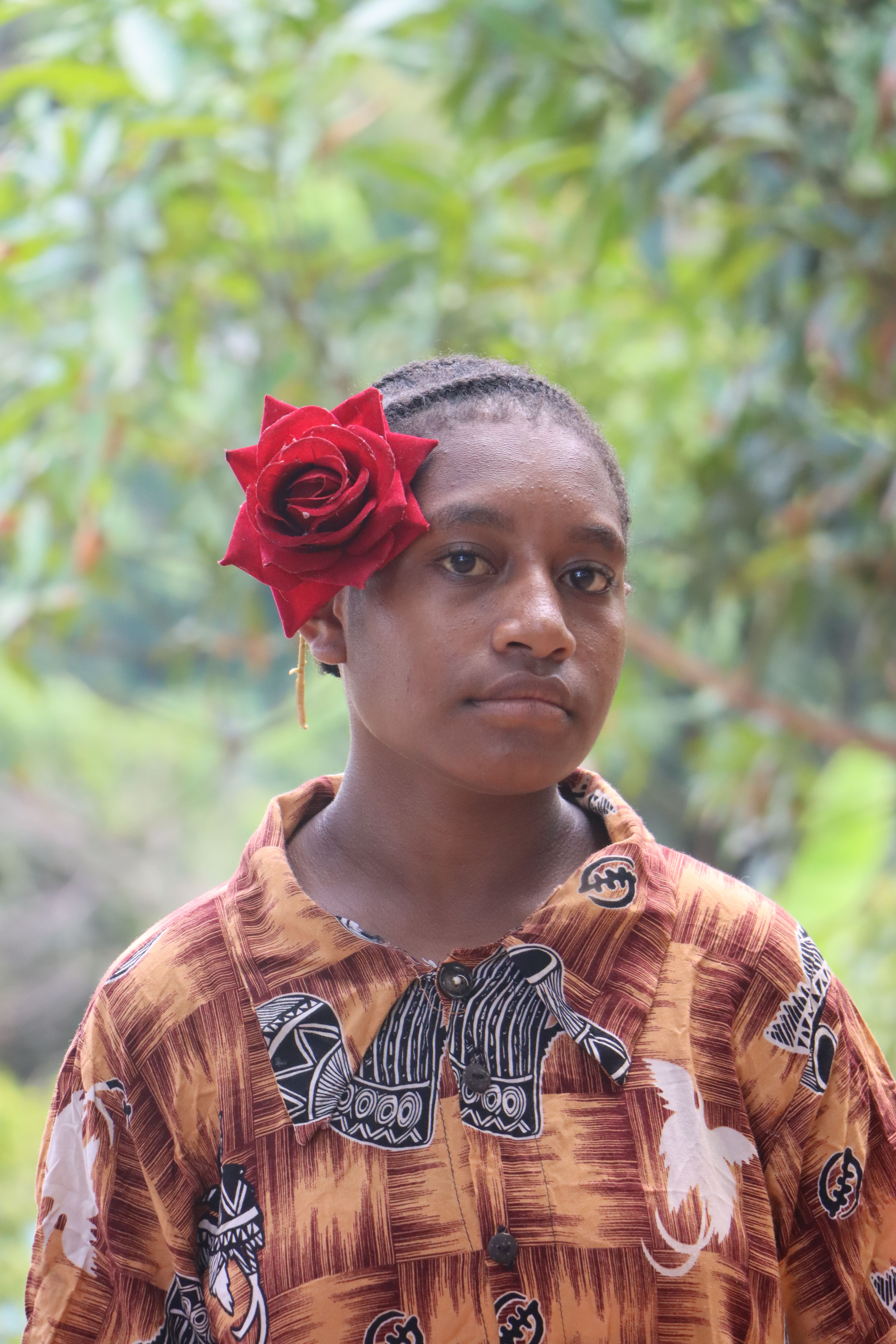 Young woman with a red rose in her hair, wearing a patterned shirt outdoors.