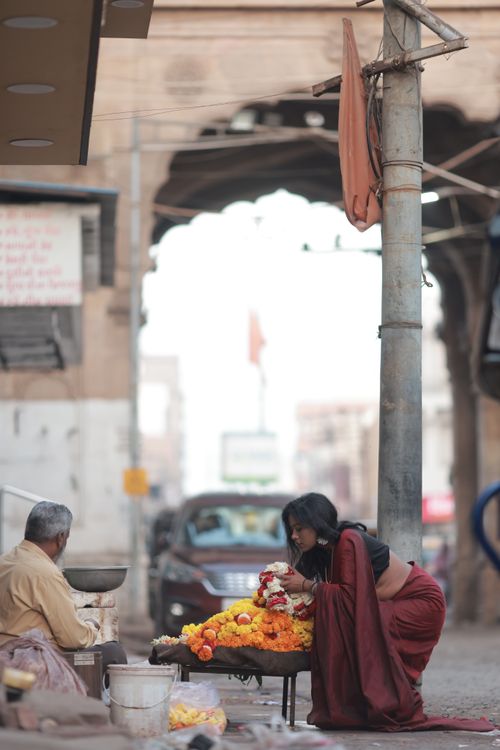 Free A woman in traditional attire arranging flowers at a bustling market stall with an archway backdrop. Stock Photo