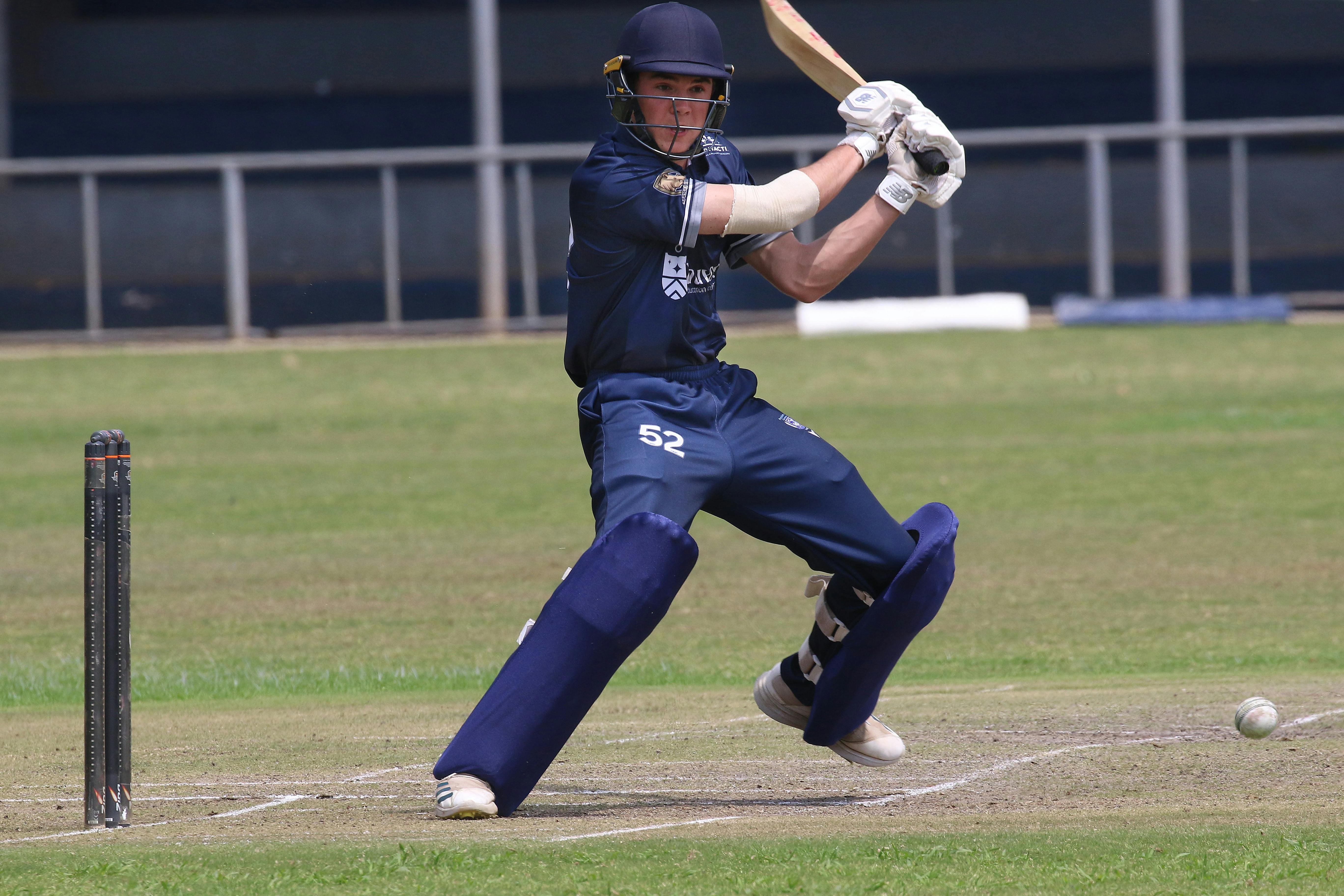 Batsman in blue uniform playing a shot on a grassy cricket field during a sunny day.