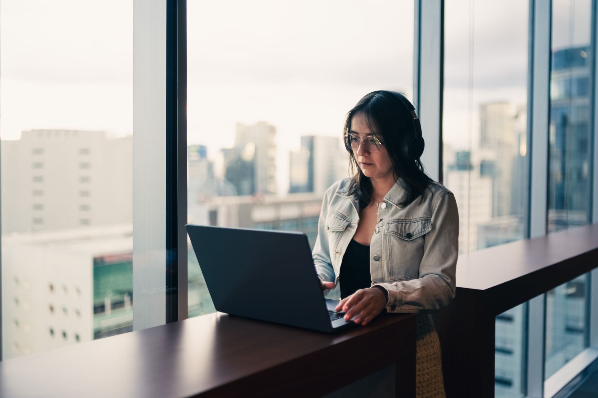 A woman using a laptop by a large window in a bright, modern office setting.