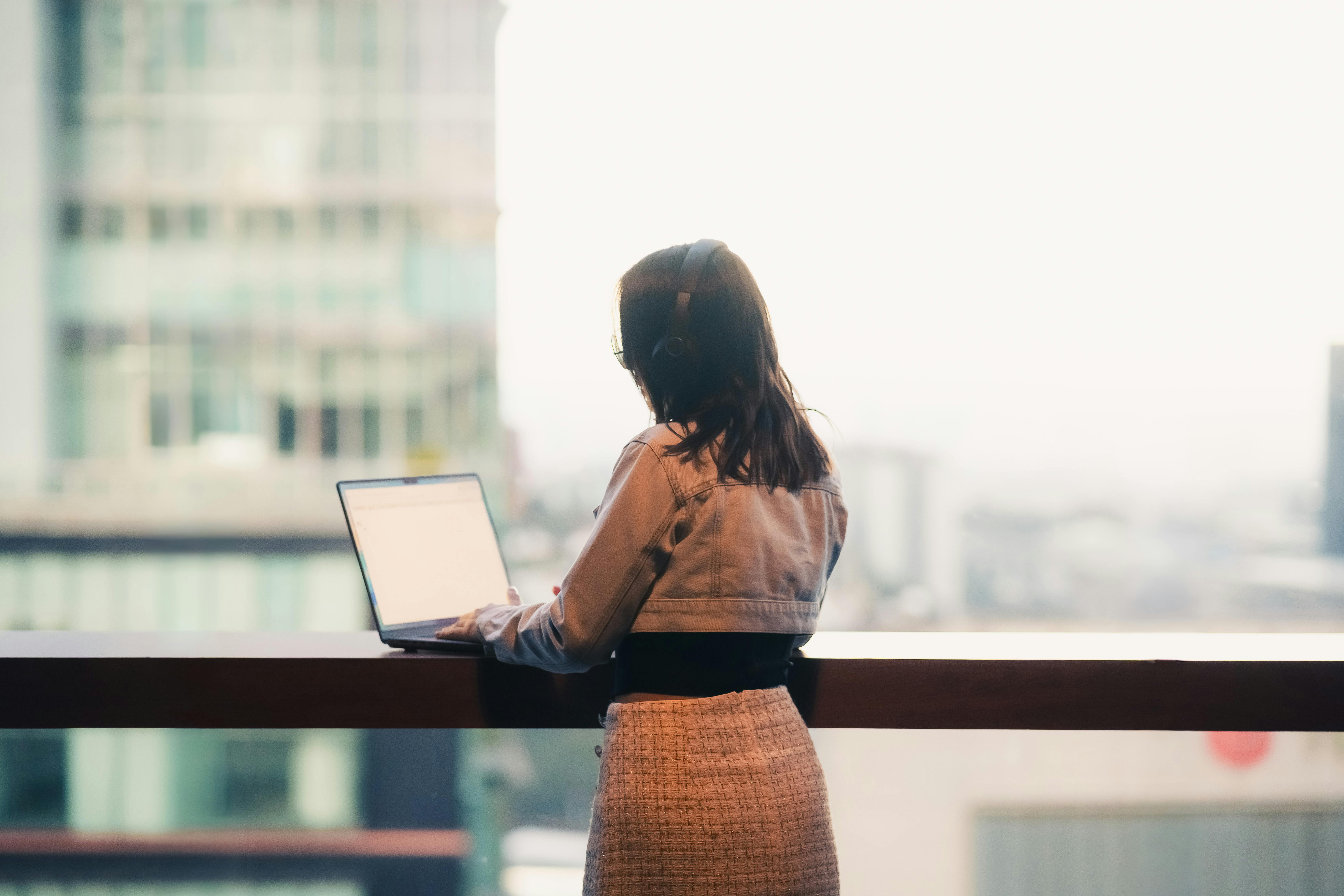 A woman wearing headphones works on her laptop overlooking a cityscape from an office or apartment balcony.
