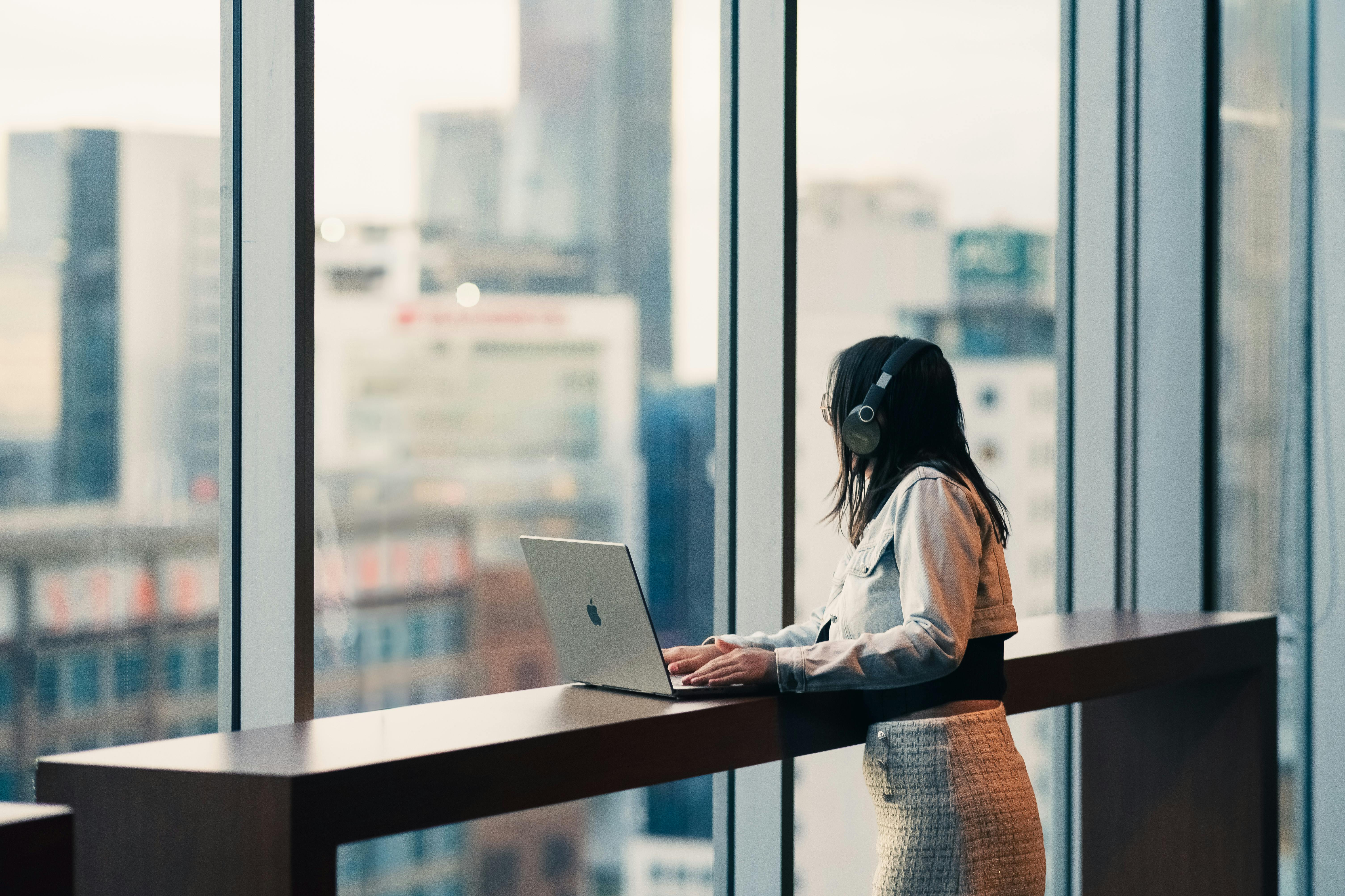 A woman wearing headphones is using a laptop in a modern office with city views.