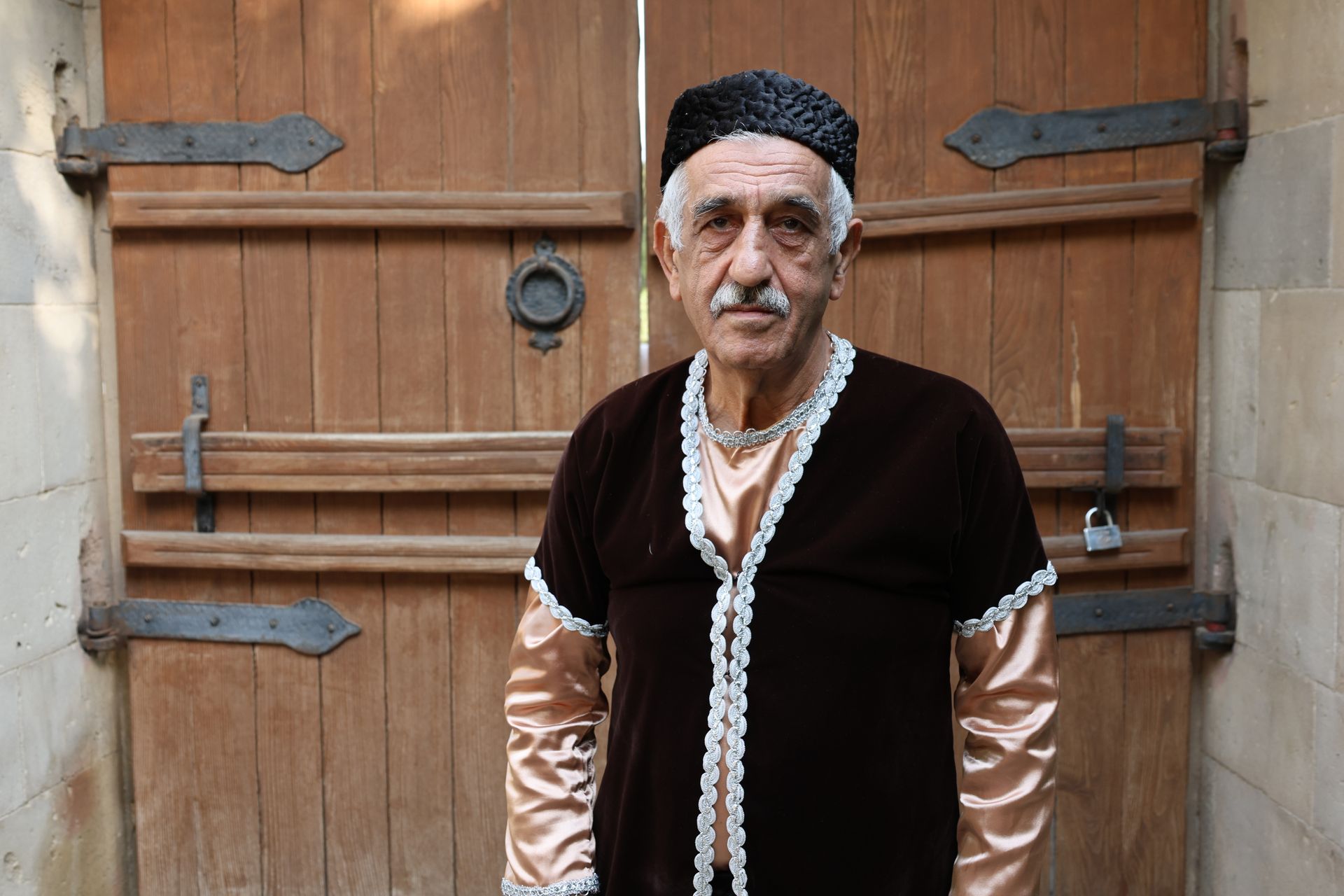 Elderly man in traditional costume stands by a wooden gate, showcasing cultural heritage.