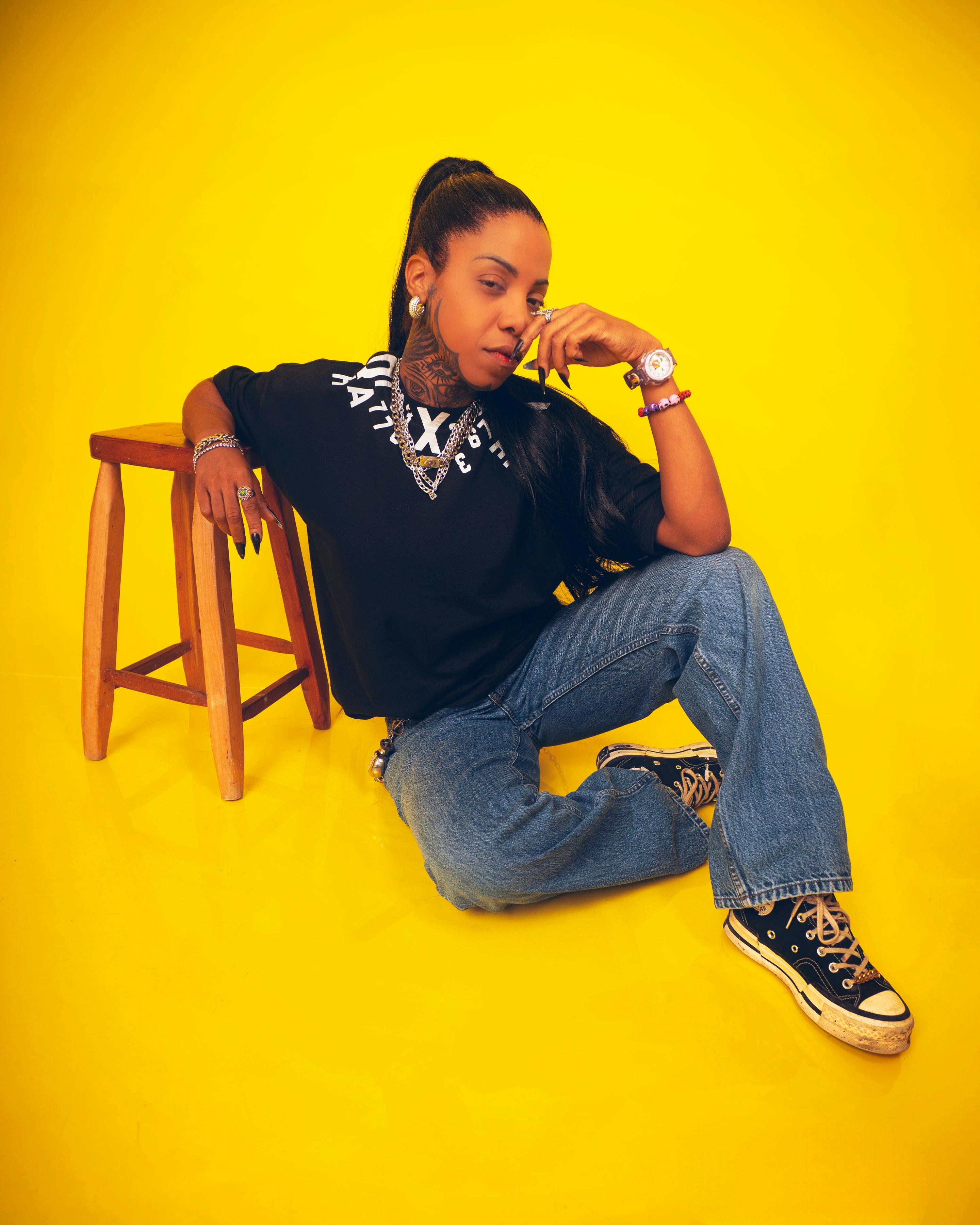 Stylish young woman in casual attire sits confidently against vibrant yellow background.