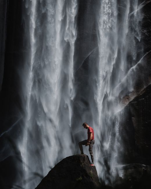 A lone hiker stands on a rock facing a powerful waterfall in Yosemite National Park.