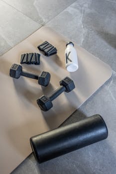 Flat lay of dumbbells, weights, and bottle on mat for fitness home workout.