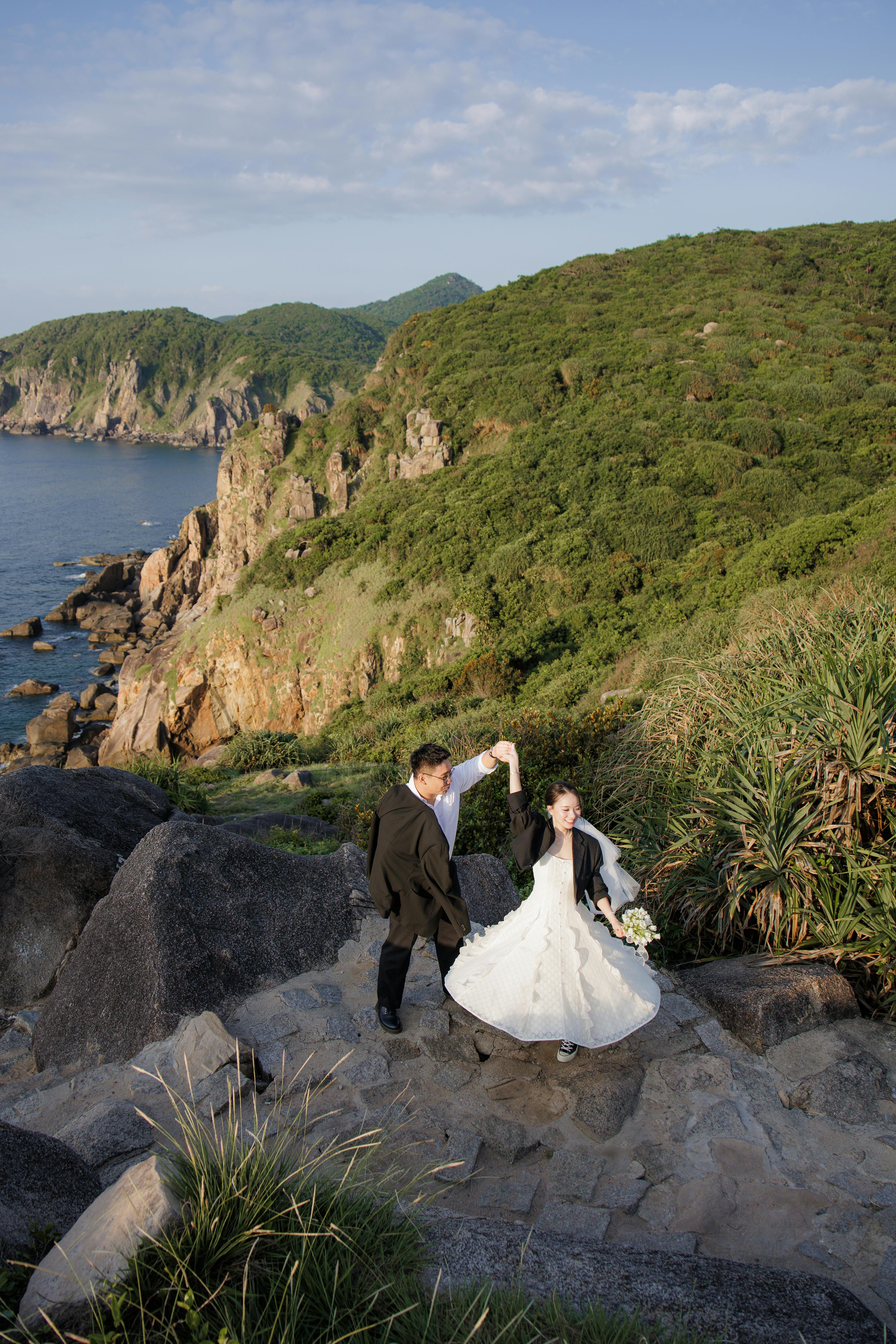 一对穿着婚纱的夫妇在风景如画的悬崖边跳舞，同时欣赏着海景和郁郁葱葱的绿化