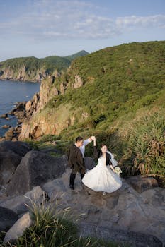 一对穿着婚纱的夫妇在风景如画的悬崖边跳舞，同时欣赏着海景和郁郁葱葱的绿化