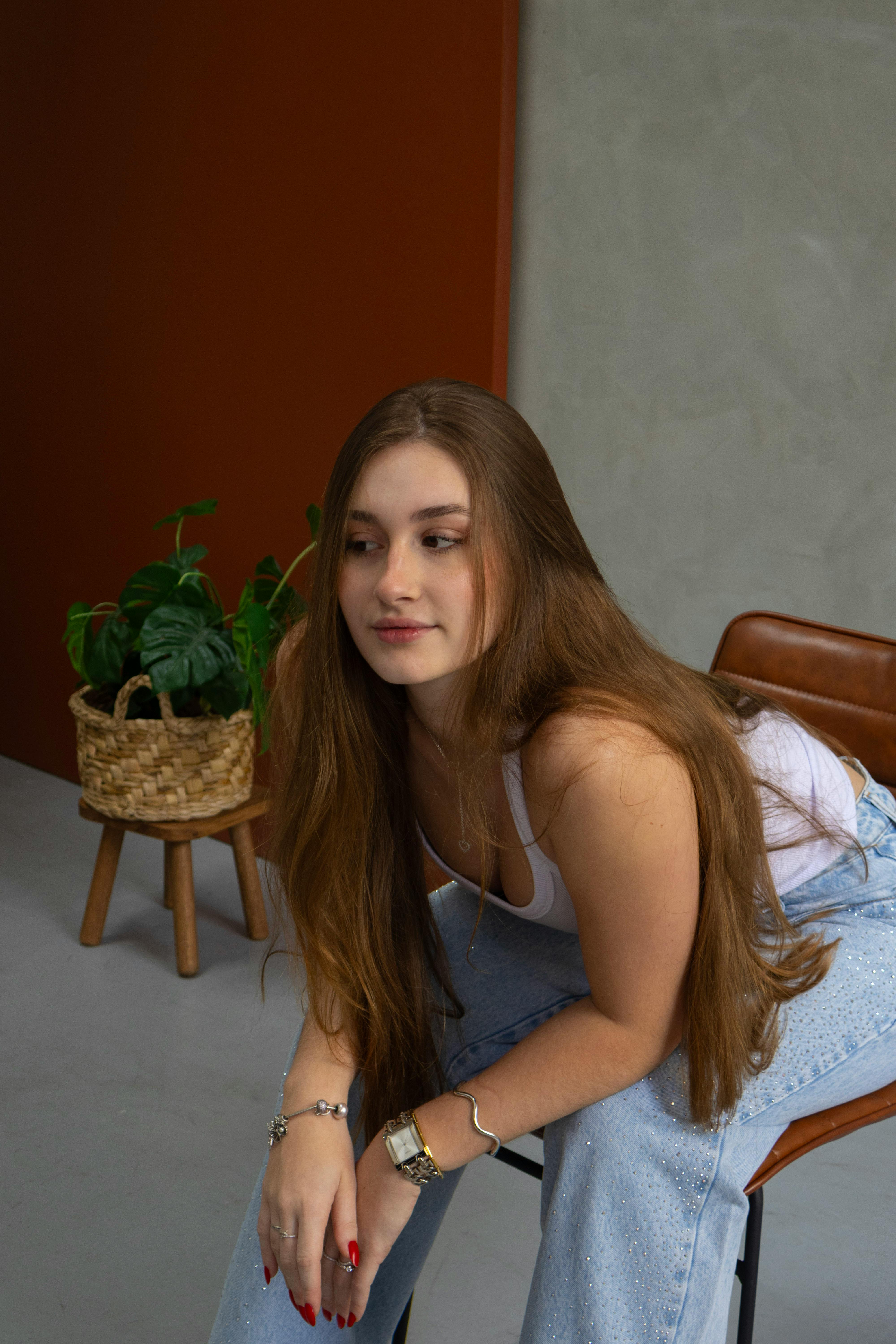 Young woman with long hair sitting indoors, wearing casual clothes with a potted plant in the background.