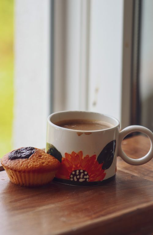 Free A cozy scene of a coffee mug with floral design and muffin on a wooden surface by a window. Stock Photo