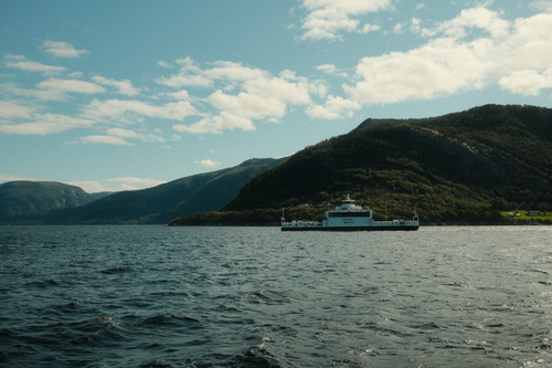 Free A serene view of a ferry crossing a fjord surrounded by lush mountains under a bright sky. Stock Photo