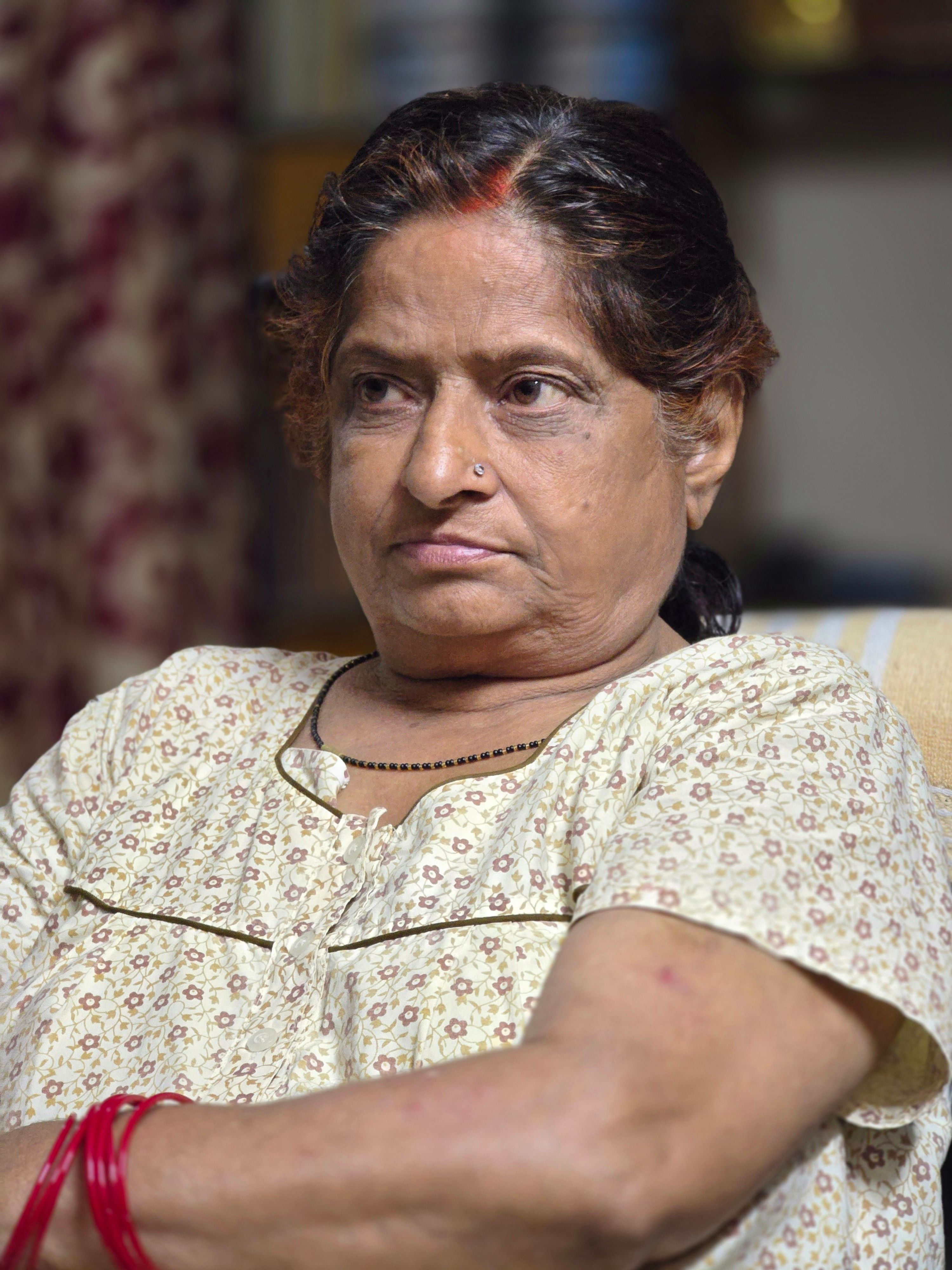 Elderly woman with a thoughtful expression sitting indoors, wearing a floral blouse.