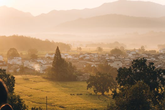 A serene sunrise over a rural village with fog covering distant hills and lush fields.