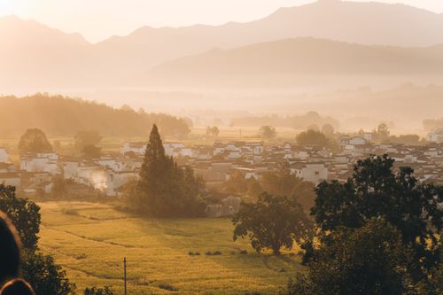 Free A serene sunrise over a rural village with fog covering distant hills and lush fields. Stock Photo