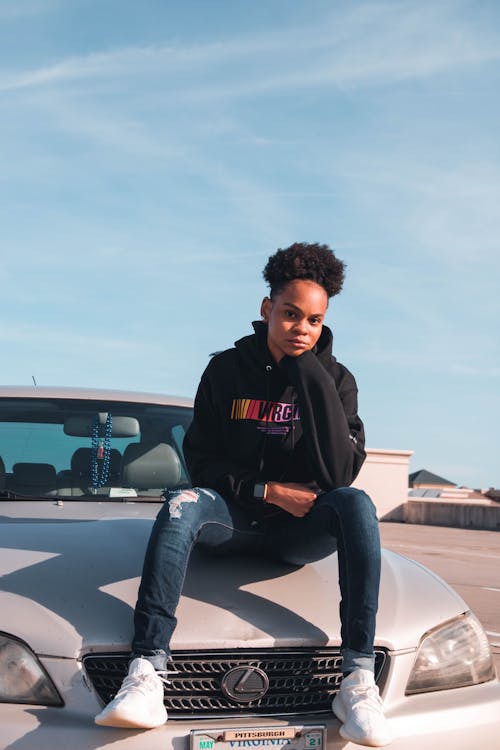 Free Person Wearing Black Hoodie and Blue Denim Jeans Sitting on Car Hood Stock Photo