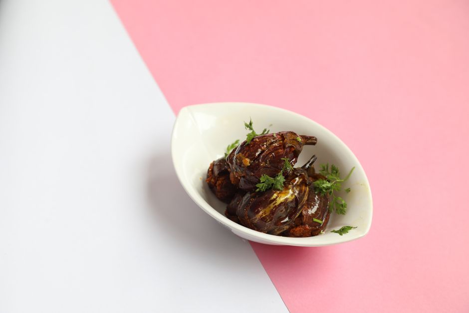 A bowl of brinjal curry with soft cubes of eggplant in a thick reddish-brown onion-tomato masala, garnished with chopped cilantro, next to a plate of steamed rice.