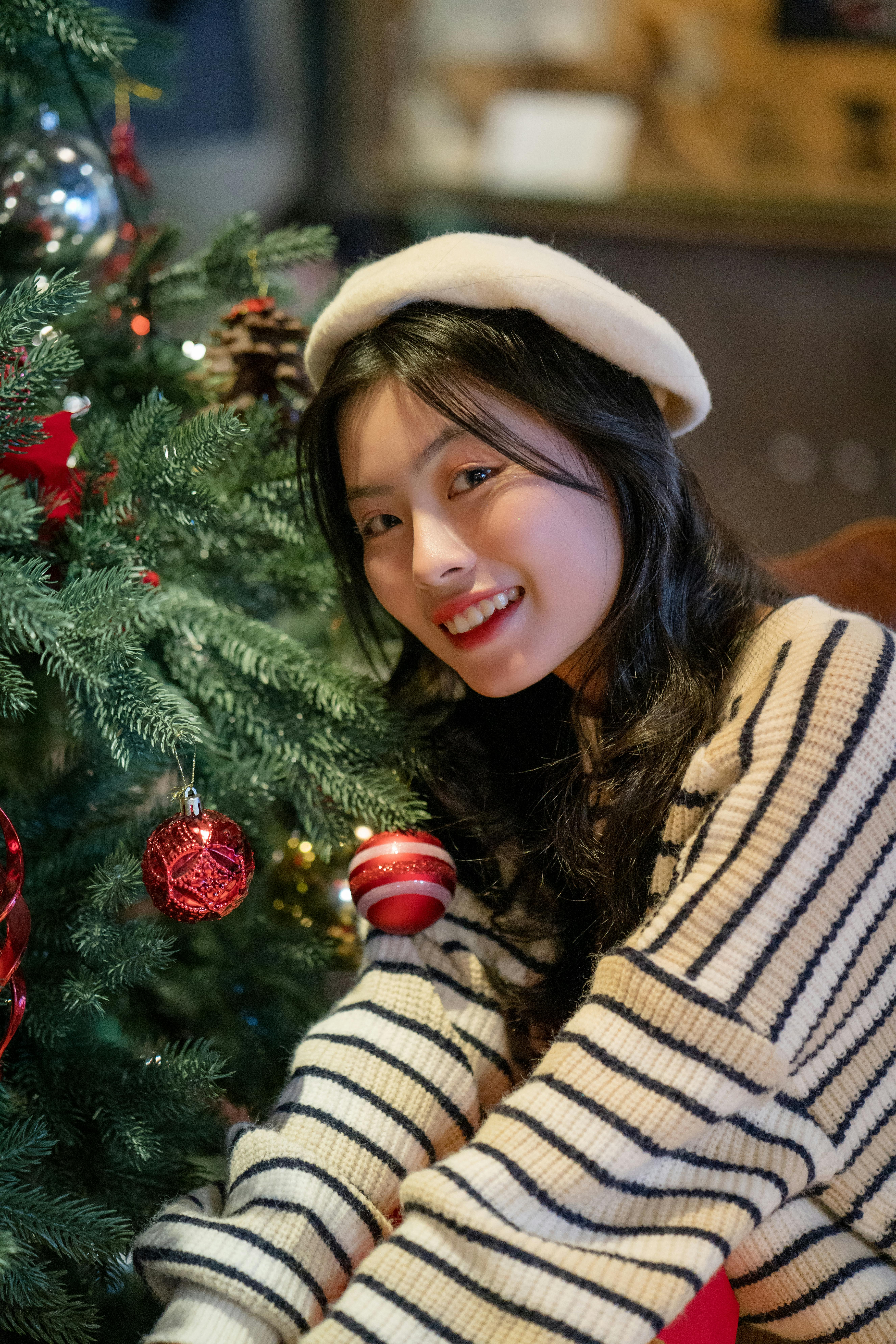 Young woman dressed warmly, smiling by a Christmas tree adorned with ornaments, exuding holiday cheer.