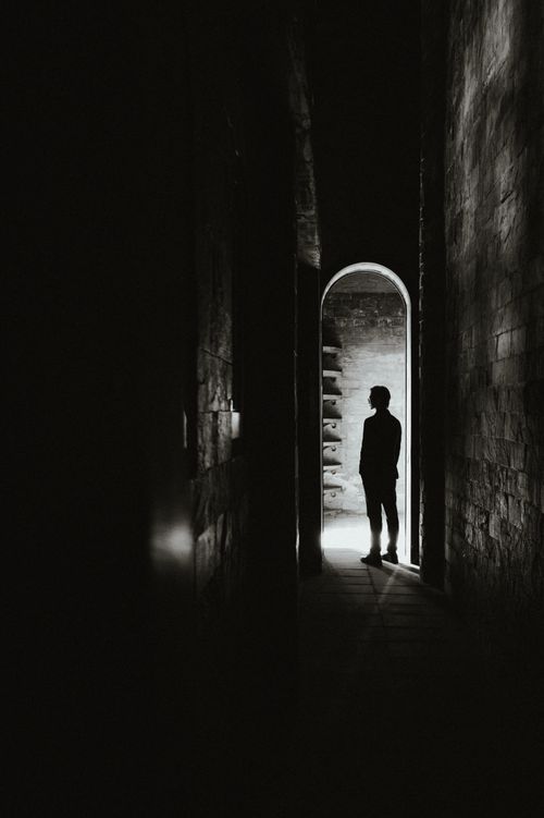 Free Silhouette of a person standing in a dark, narrow hallway with a light source at the end. Stock Photo