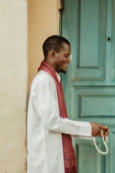 Side view of a smiling man in traditional attire with a rope, standing by a teal door.