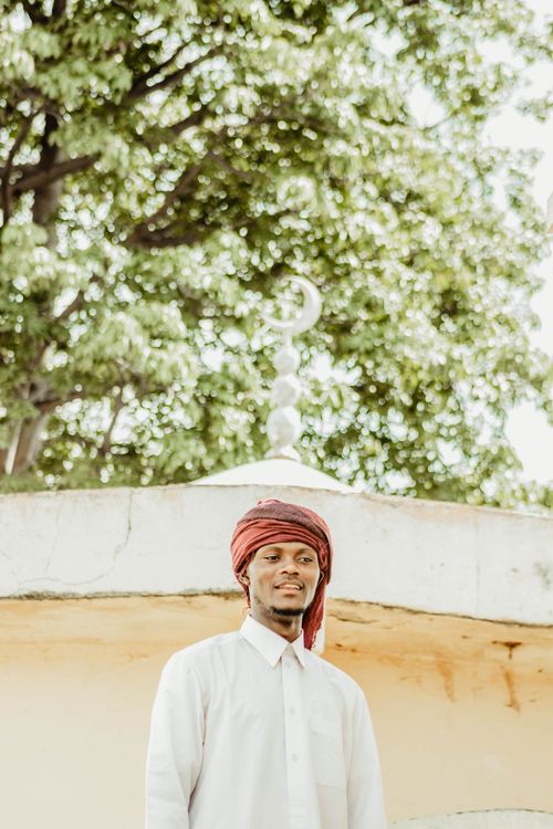 Free A man wearing traditional clothing stands under a crescent moon ornament, surrounded by lush greenery. Stock Photo