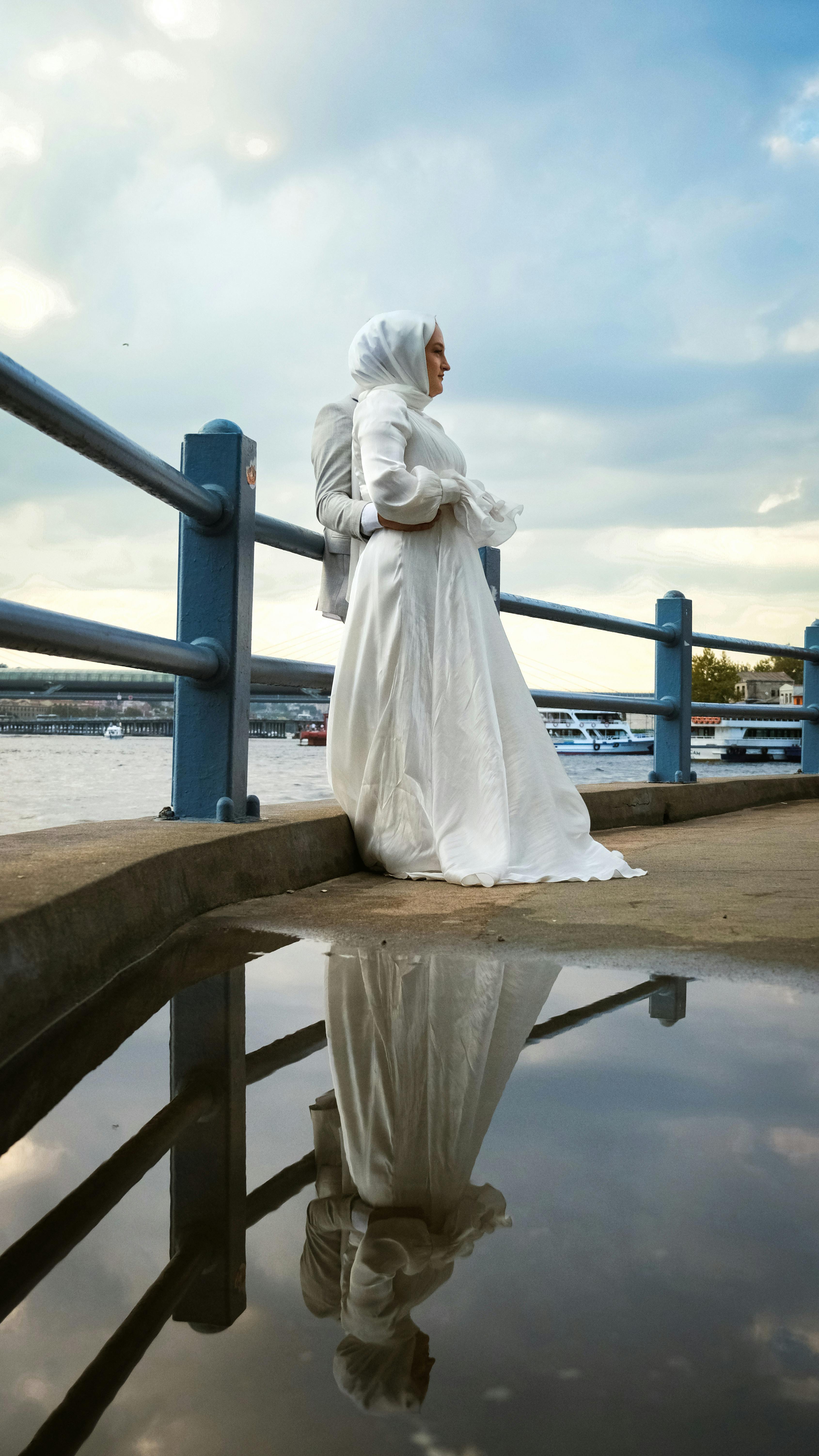 身穿白色礼服的新娘站在河边，她的倒影映在水中