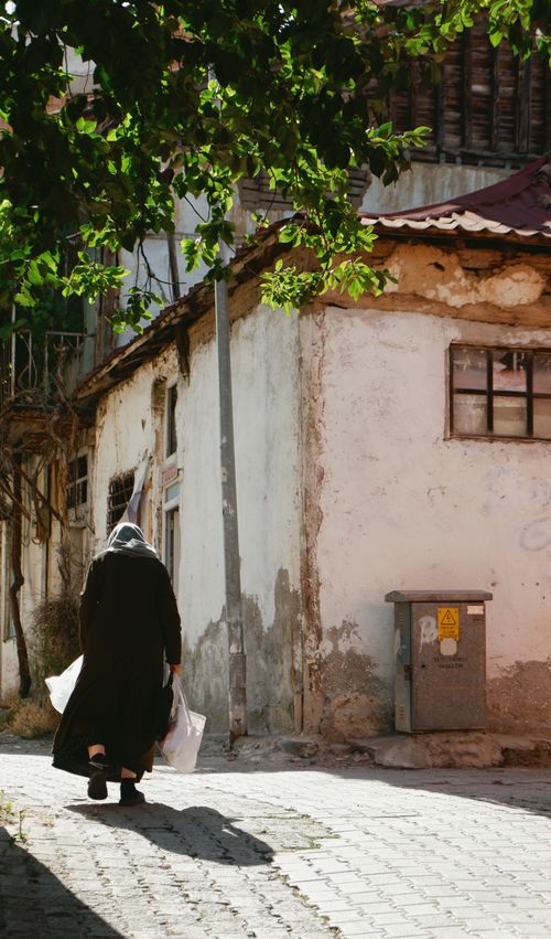 Free Back view of an elderly woman walking down a sunlit, historic alley carrying shopping bags. Stock Photo