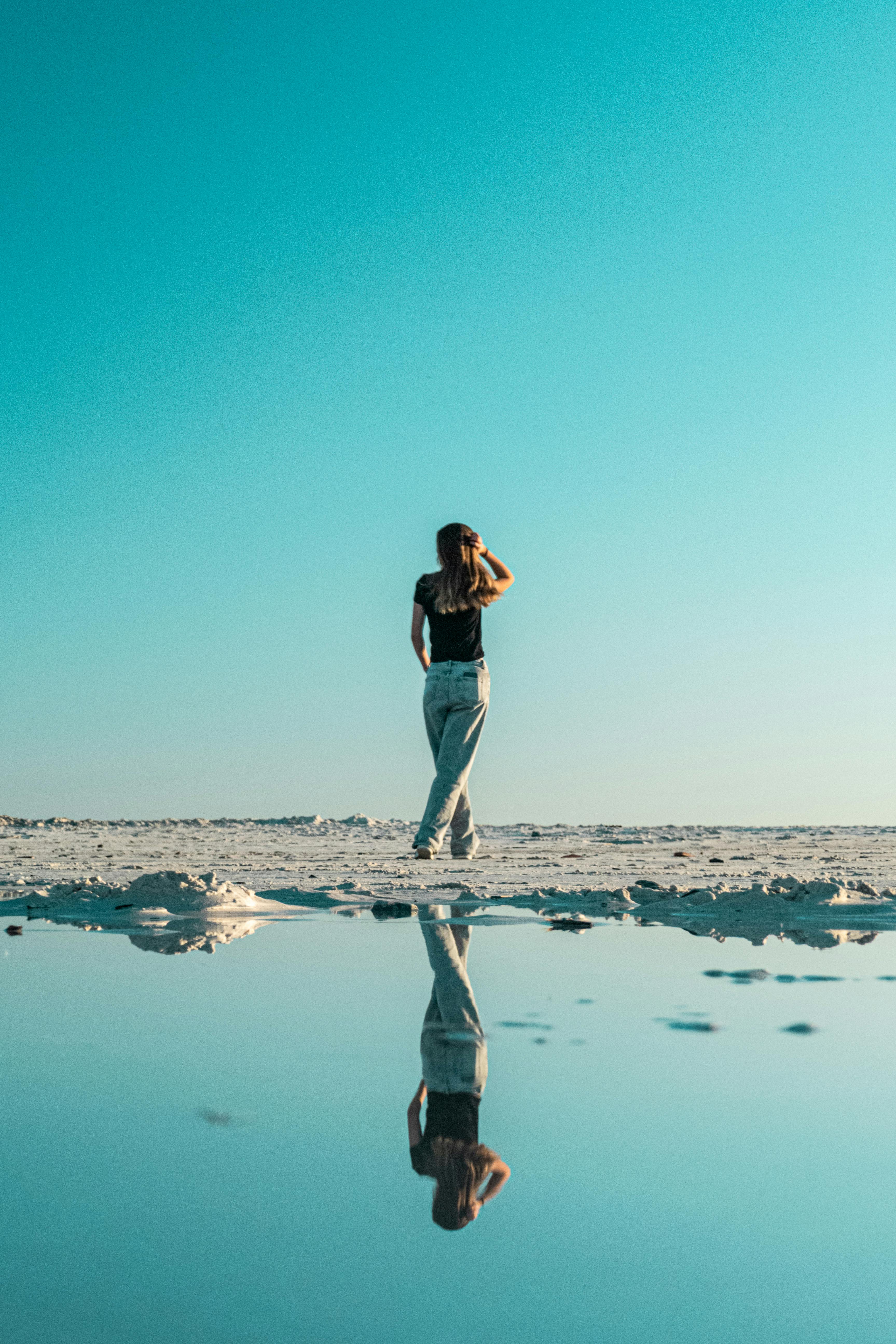 一名女子站在宁静的海滩上，清澈的蓝天下平静的水中倒影