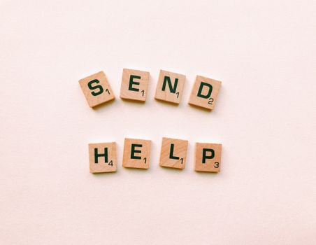 Wooden Scrabble tiles arranged to form the phrase 'send help' on a light pink background.