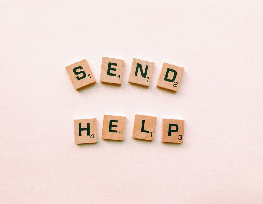 Wooden Scrabble tiles arranged to form the phrase 'send help' on a light pink background.