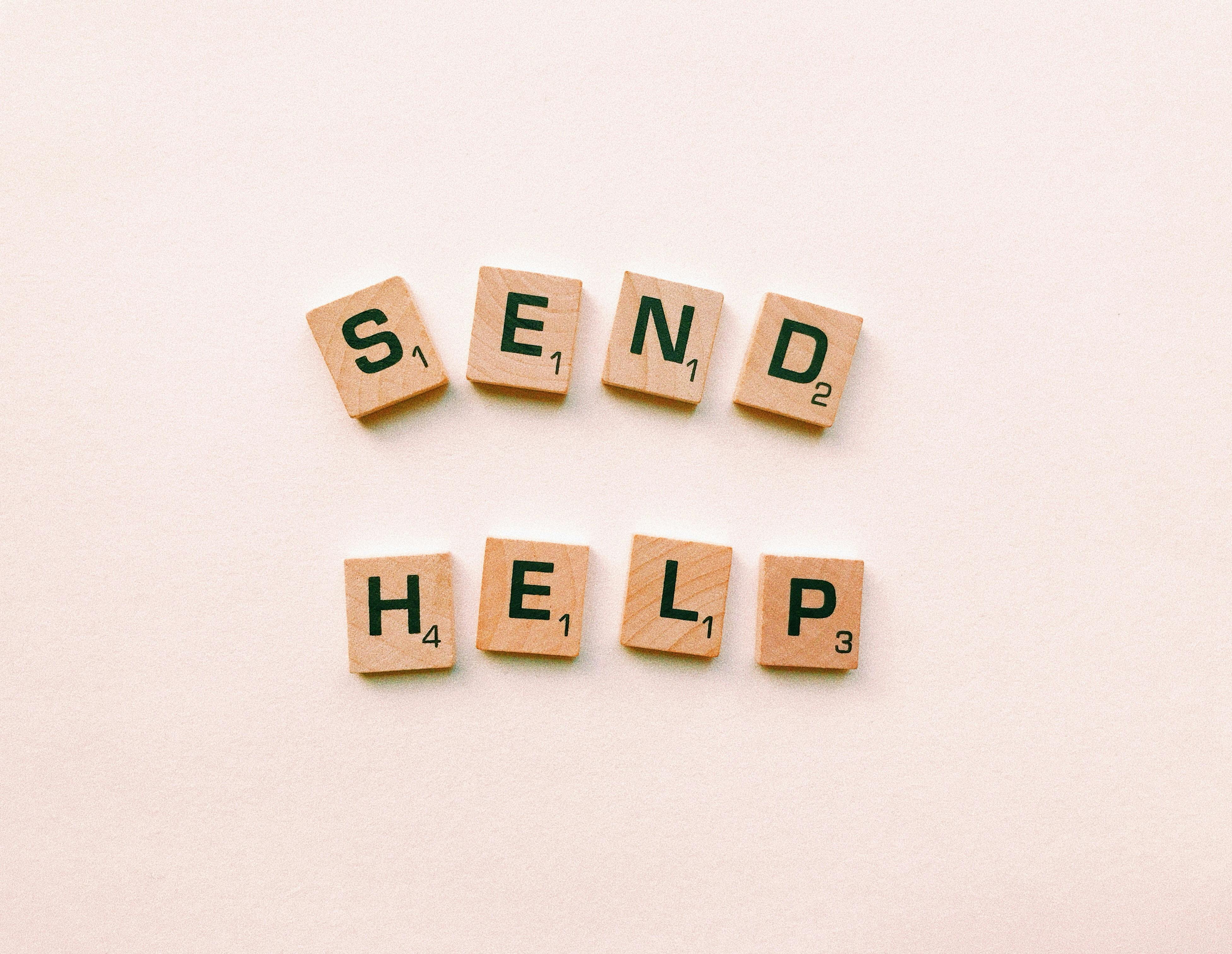 Free Wooden Scrabble tiles arranged to form the phrase 'send help' on a light pink background. Stock Photo