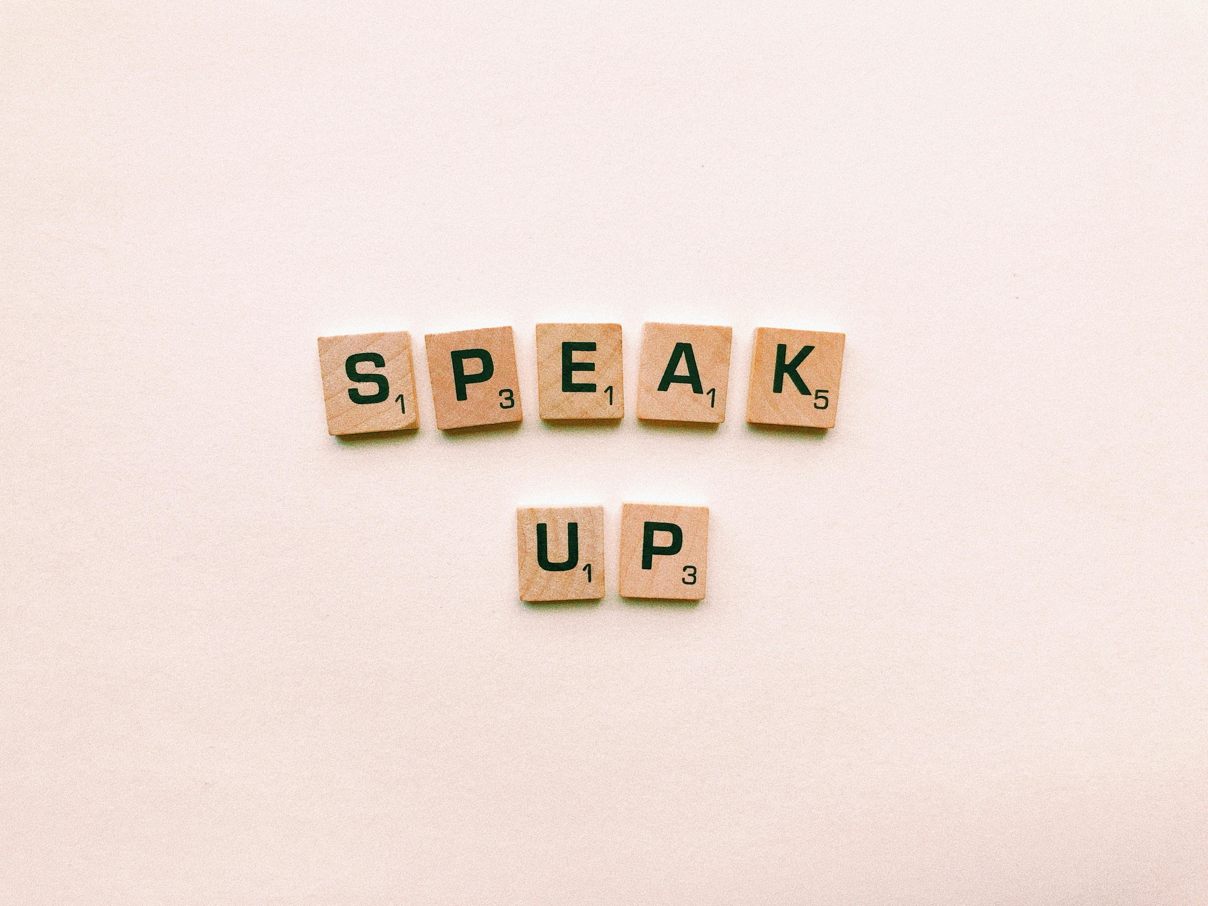 Wooden Scrabble tiles spelling 'Speak Up' on a pink background.