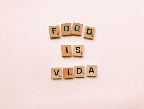Wooden letter tiles spell 'FOOD IS VIDA' on a soft pink background.