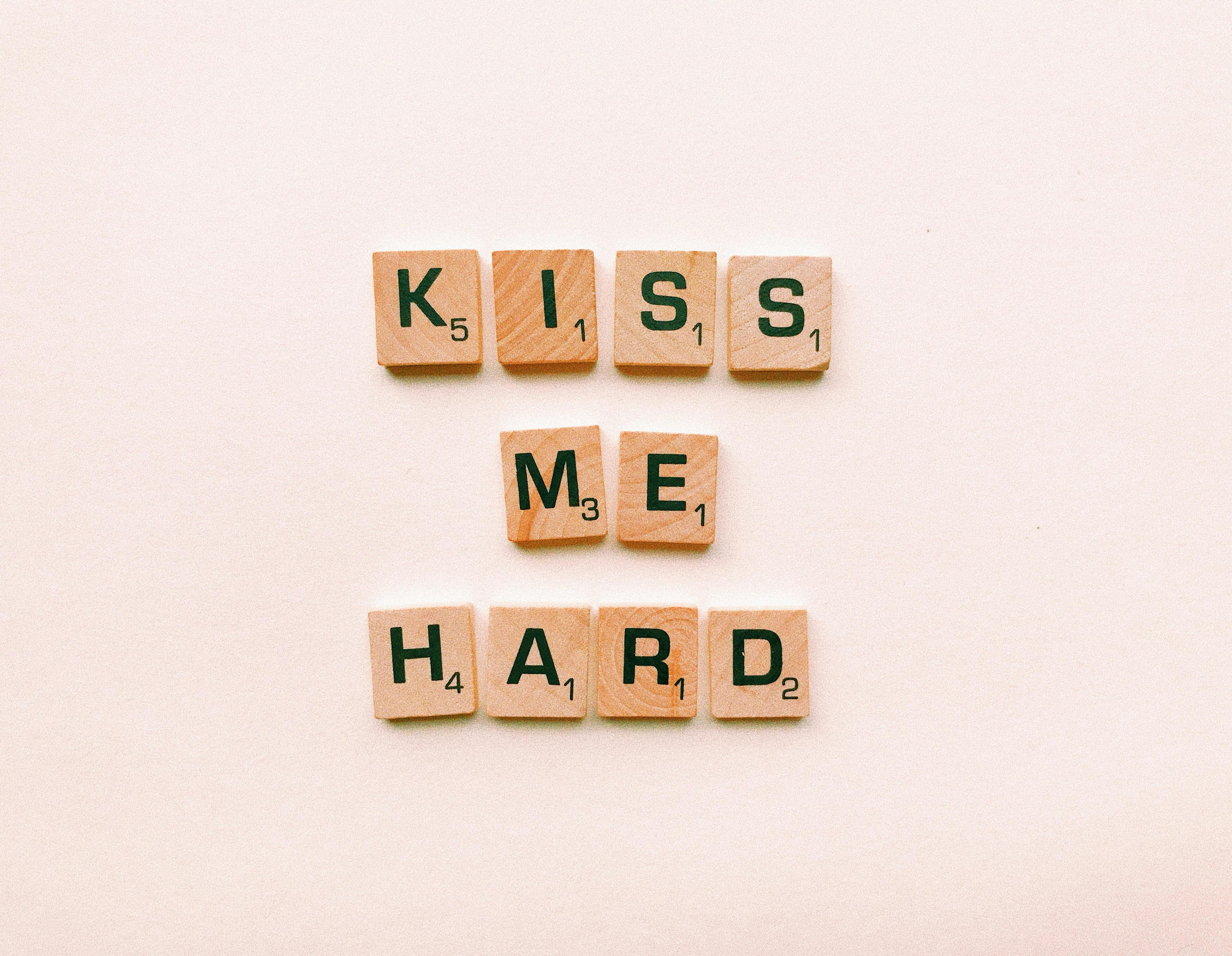 Wooden Scrabble tiles spell out 'Kiss Me Hard' against a pink background. Perfect for romantic themes.