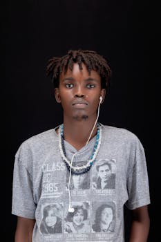 Portrait of a young man with dreadlocks, wearing a graphic t-shirt and earphones against a black background.