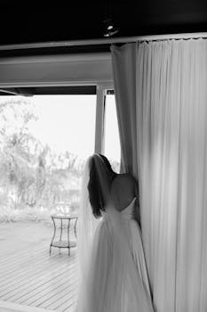 一位穿着婚纱的新娘透过窗帘偷看，捕捉到婚礼前的一个宁静时刻