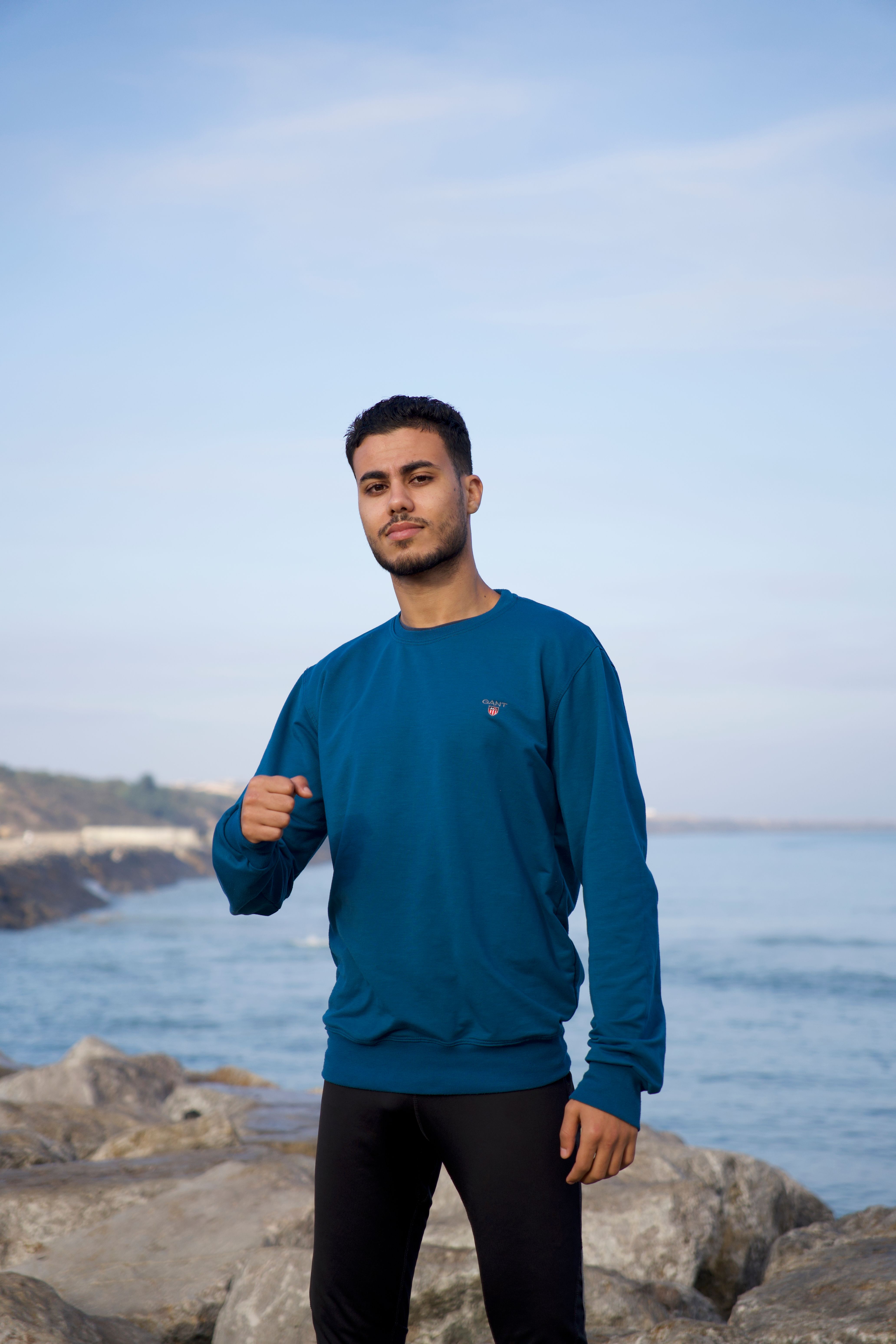 Young man in blue sweater standing on rocky beach under clear sky.
