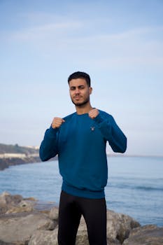 自信的年轻人穿着蓝色毛衣和锻炼装备，在岩石海岸上摆姿势