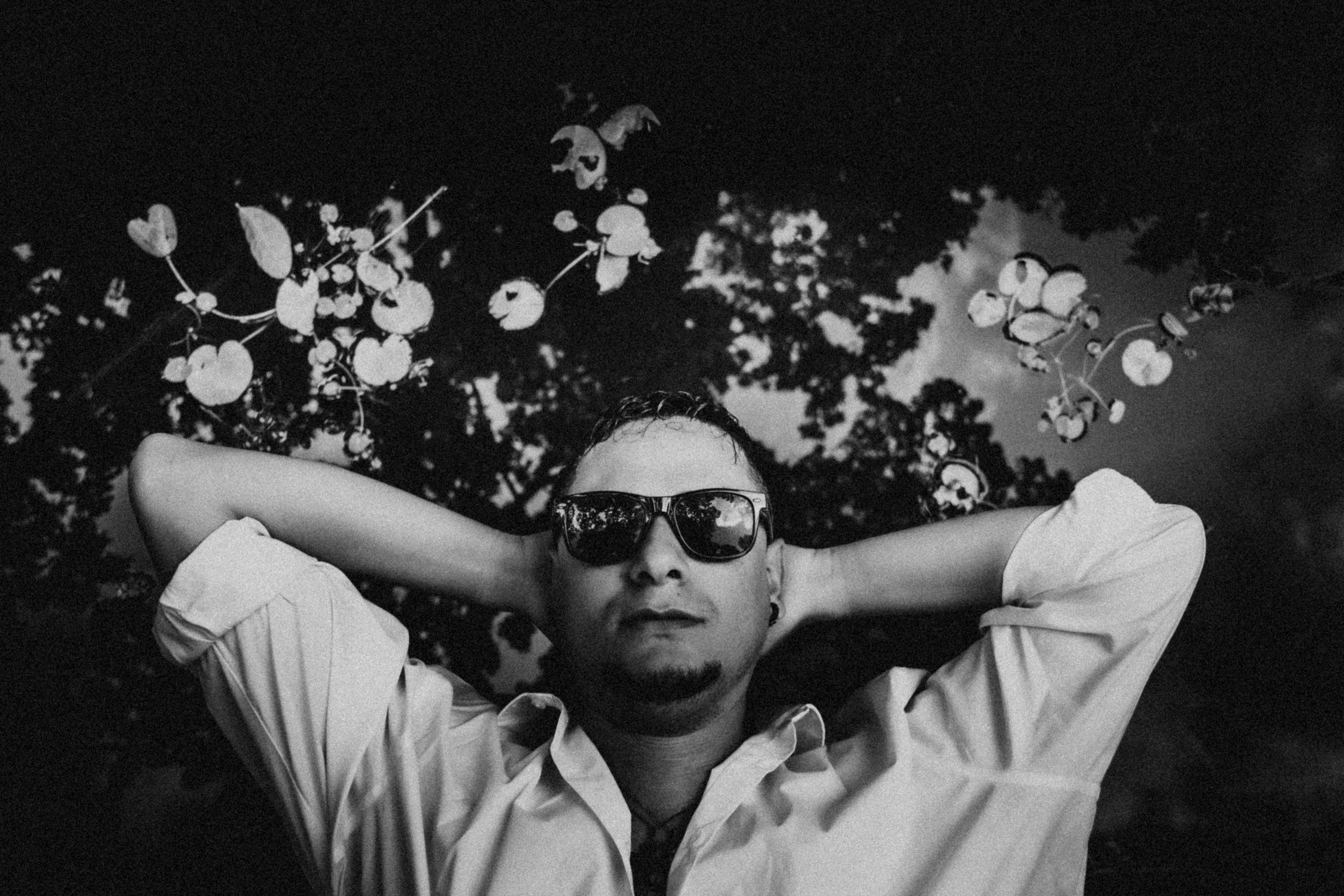 A monochrome portrait of a man wearing sunglasses, relaxing under tree shade.