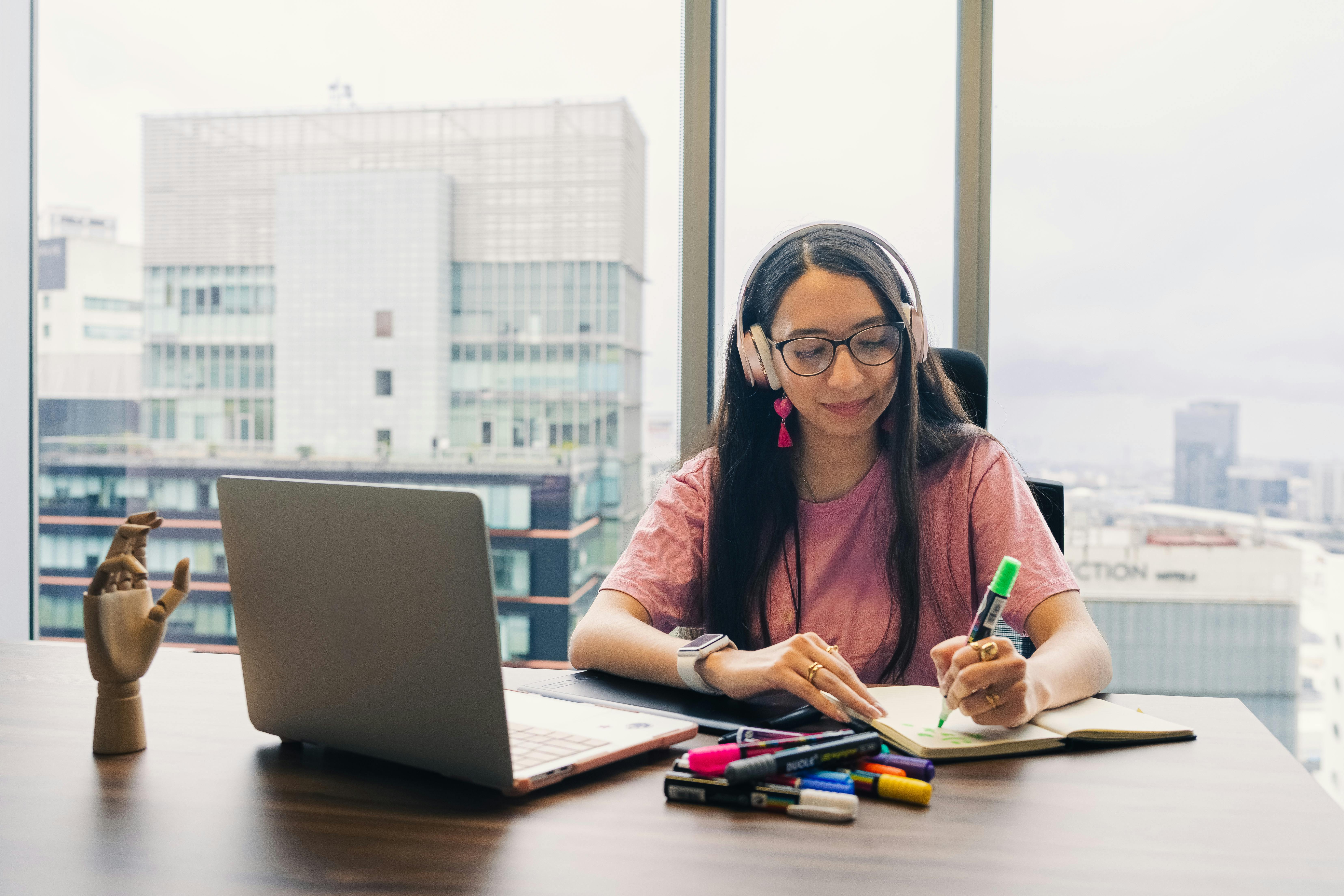 Young woman wearing headphones, writing in notebook at office desk with city view.