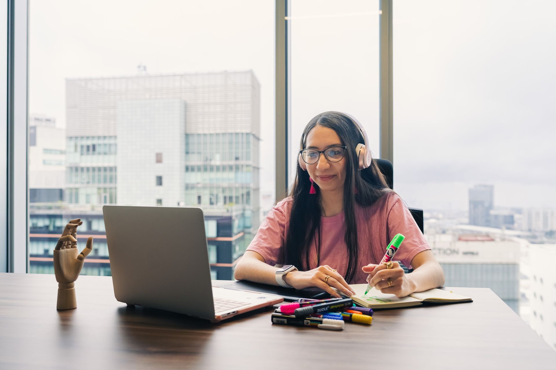 Woman wearing headphones working on a laptop in a modern office with a city view.
