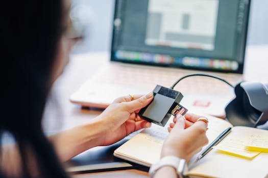 A person inserts an SD card into a reader next to a laptop on a desk.