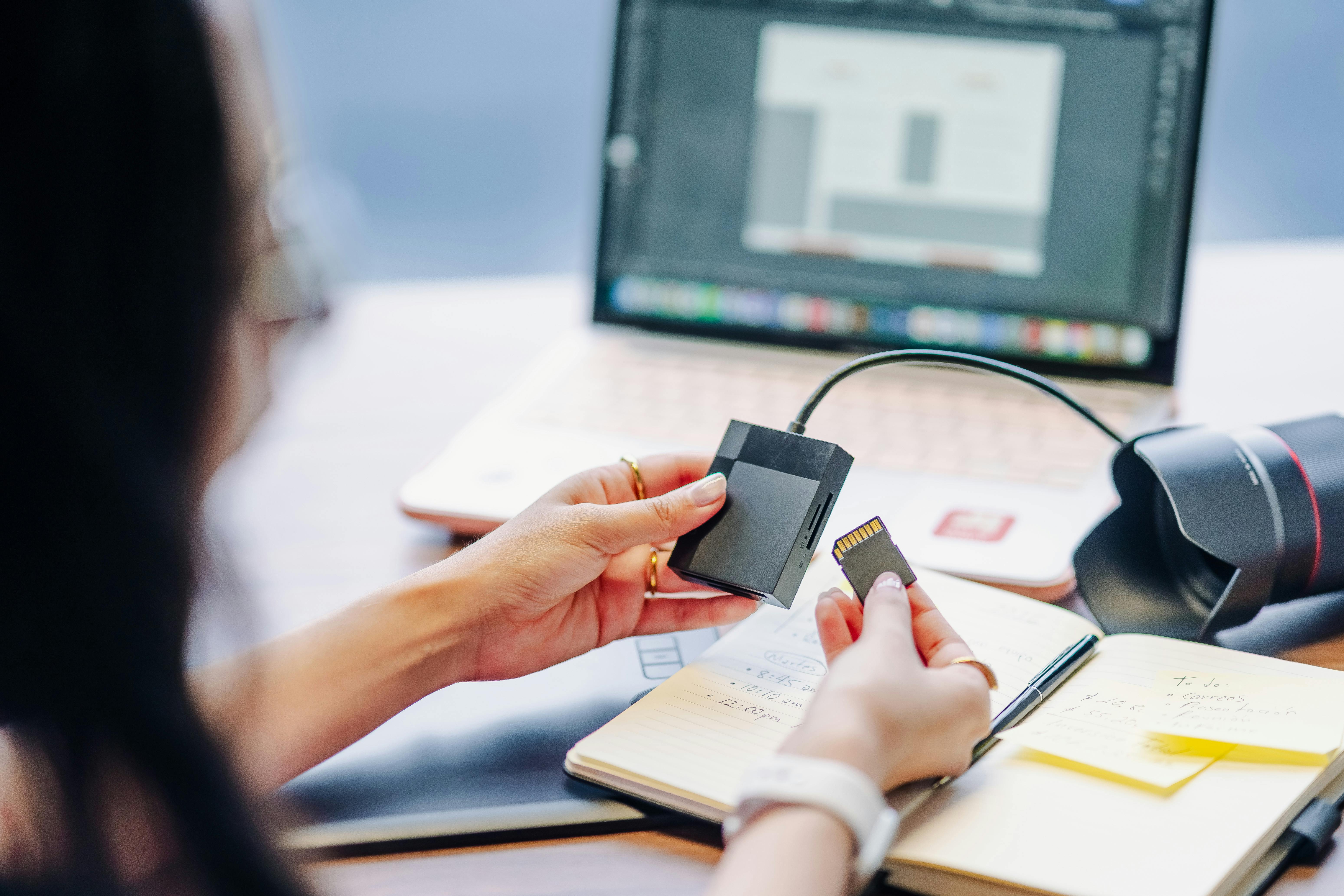 A person prepares to edit photos on a laptop by inserting an SD card, with camera gear on the desk.