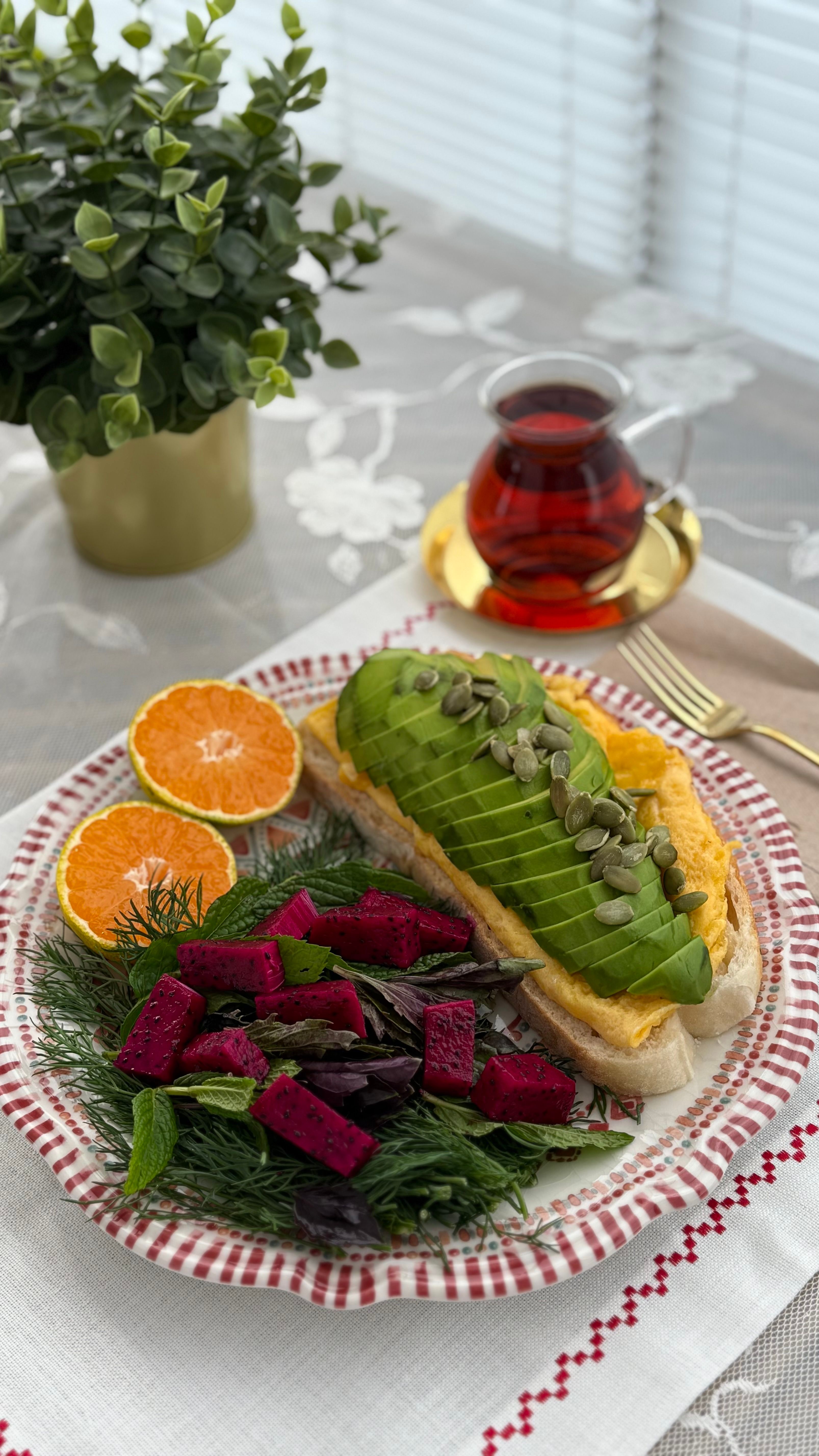 A vibrant, healthy breakfast plate featuring avocado toast, dragon fruit salad, orange slices, and tea.